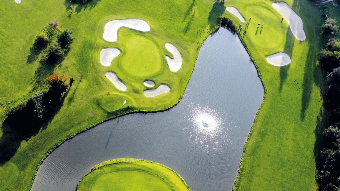 <p>Eine malerische Golfanlage mit grünen Fairways und Sandbunkern. Ein ruhiger Teich spiegelt die Sonne wider.</p> | © GEPA-pictures - Murhof Gruppe