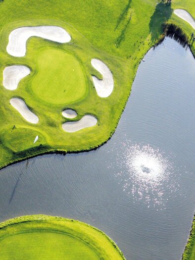 <p>Eine malerische Golfanlage mit grünen Fairways und Sandbunkern. Ein ruhiger Teich spiegelt die Sonne wider.</p> | © GEPA-pictures - Murhof Gruppe