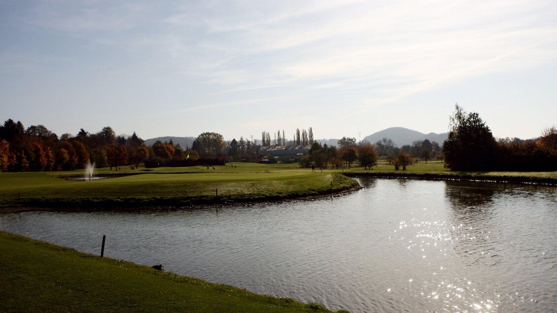 <p>Eine gemütliche Golfanlage mit herbstlichen Bäumen im Hintergrund. Drei Personen spielen Golf am Ufer eines ruhigen Gewässers.</p>
 | © GEPA-pictures - Murhof Gruppe
