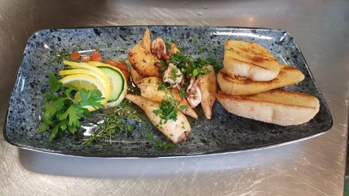 Ein Teller mit gegrilltem Tintenfisch, garniert mit frischen Kräutern und Zitrone. Dazu gibt es einige Scheiben geröstetes Brot. | © Gotthardt's Gusto