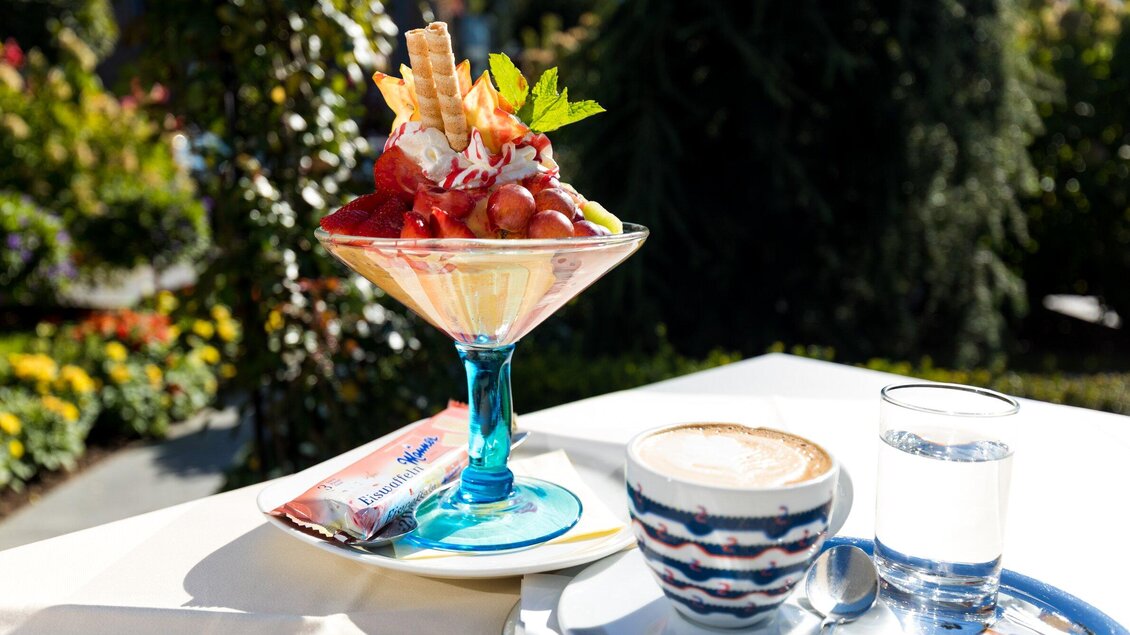 Ein leckeres Eisbecher mit frischen Früchten und Sahne, serviert in einem schönen Glas. Neben dem Eis steht eine Tasse Kaffee und ein Glas Wasser. | © TV Region Graz - Harry Schiffer