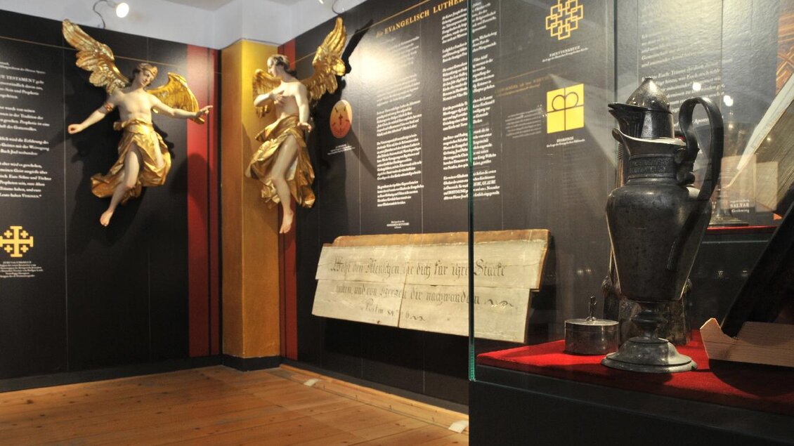 Ein Ausstellungsraum mit goldenen Engelstatuen und historischen Objekten. Auf der Wand befinden sich informative Texte und eine große Tafel. | © Gröbming Museum