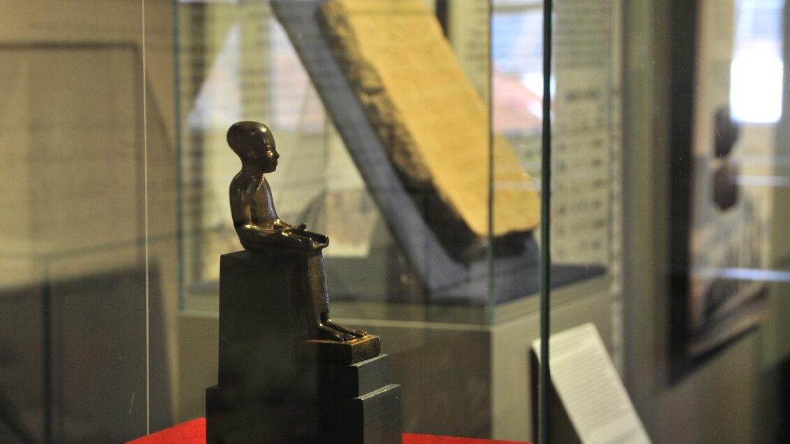 Eine kleine bronze Statue eines sitzenden Kindes auf einem Podest in einem Vitrinenraum. Im Hintergrund sind Texte und andere historische Objekte zu sehen. | © Gröbming Museum