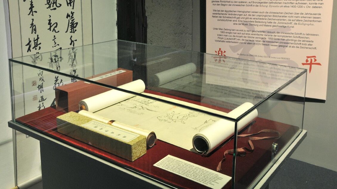 Ein Vitrinenexponat mit alten Schriftrollen und einer Zeichnung. Im Hintergrund sind chinesische Schriftzeichen zu sehen. | © Gröbming Museum