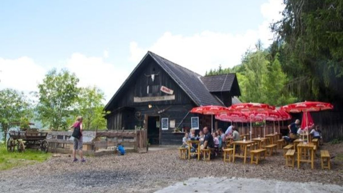 Ein gemütliches Restaurant in den Bergen mit Holzgebäude und stilvollen Terrassen. Es gibt mehrere Tische unter roten Sonnenschirmen, die Gäste genießen die Atmosphäre. | © Gröbminger Alm