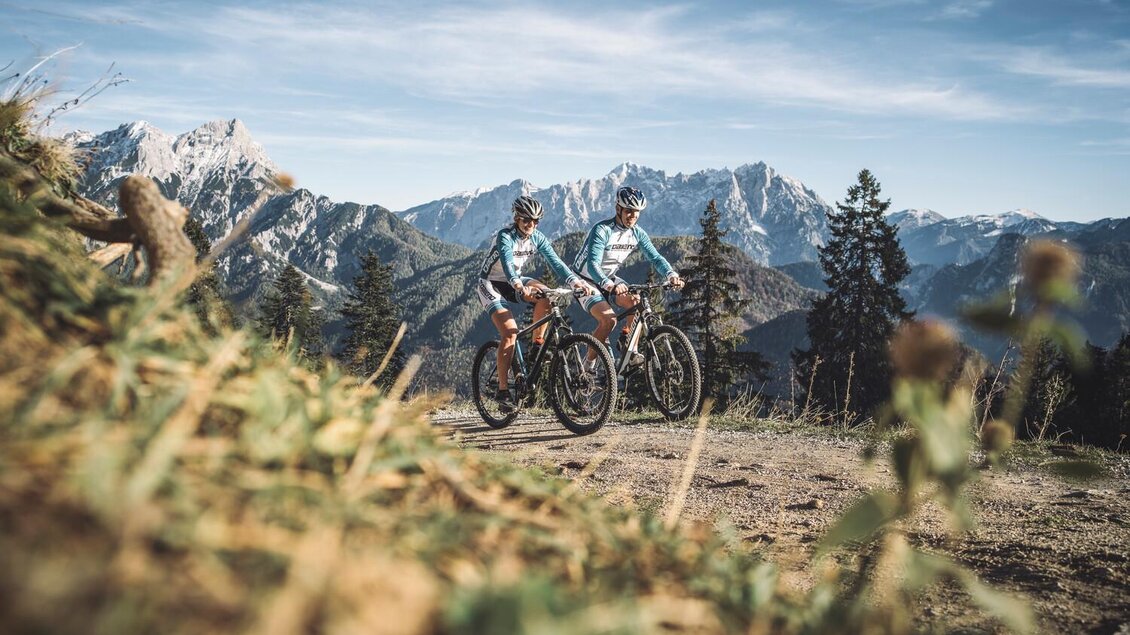 Zwei Radfahrer stehen auf einem Berghügel mit atemberaubendem Blick auf die umliegenden Berge. Der Himmel ist klar und die Landschaft ist von grünen Pflanzen und Bäumen umgeben. | © Stefan Leitner