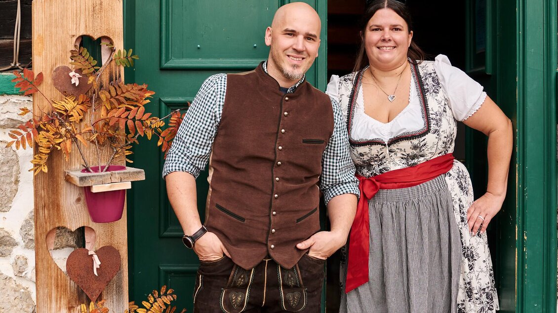 Ein Paar in traditioneller bayerischer Kleidung steht vor einer grünen Tür. Im Hintergrund sind herbstliche Dekorationen zu sehen.