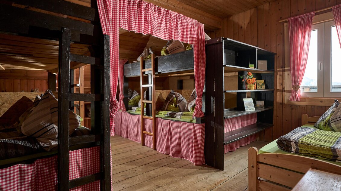 Ein gemütlicher Schlafsaal mit Etagenbetten und Holzverkleidung. Die Fenster sind mit rosa Vorhängen geschmückt, und die Betten sind einladend mit Kissen und Decken ausgestattet.