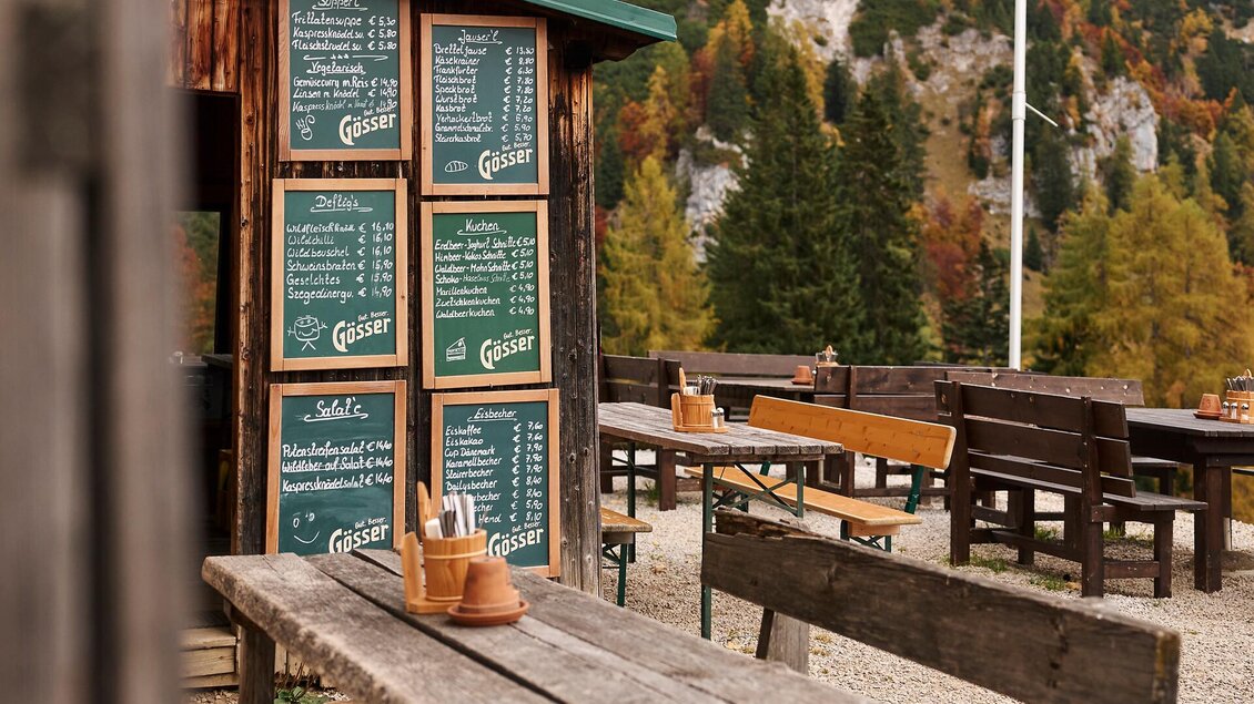 Eine rustikale Außenterrasse mit Holztischen und -bänken. Im Hintergrund befindet sich ein Schaukasten mit der Speisekarte.