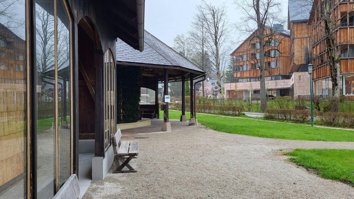 Eine ruhige Umgebung mit einem Holzpavillon und einem Weg, gesäumt von Bäumen. Im Hintergrund sieht man moderne Gebäude. | © Petra Kirchschlager