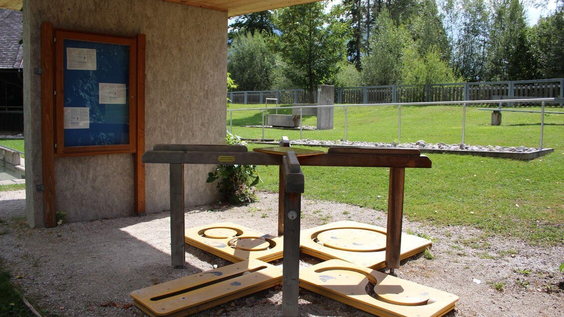 Ein Spielplatz mit einem Holzspielgerät und einer Infotafel. Der Bereich ist von grünem Gras und Bäumen umgeben. | © Viola Lechner