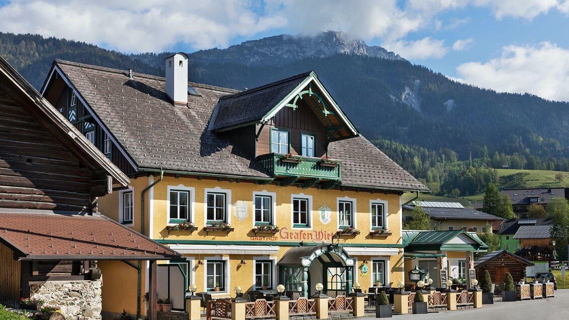 Ein malerisches Gasthaus mit gelber Fassade und grünem Balkongeländer inmitten einer bergigen Landschaft. Die Umgebung ist von sanften Hügeln und klaren, blauen Himmel umgeben. | © GrafenWirt