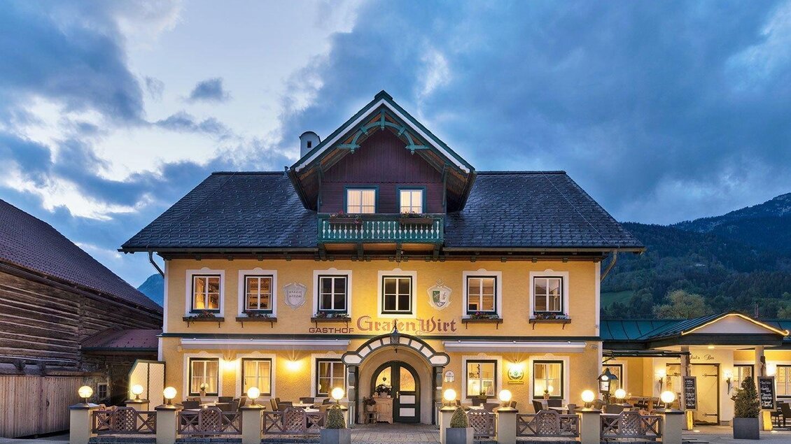 Ein charmantes Gasthaus mit gelber Fassade und doppelter Dachlinie. Der Himmel ist bewölkt und es ist Abenddämmerung. | ©  GrafenWirt 