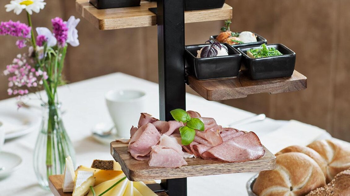 Ein stilvolles Frühstücksarrangement mit verschiedenen Aufschnittsorten, Käse, frischen Früchten und Marmelade. Dazu sind Brötchen und Blumen dekorativ angerichtet. | © GrafenWirt