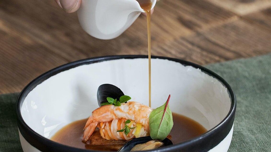 Ein Hand gießt Brühe über eine Garnelenspeise in einer eleganten Schale. Dazu sind frische Kräuter und eine dunkle Unterlage zu sehen. | © GrafenWirt 