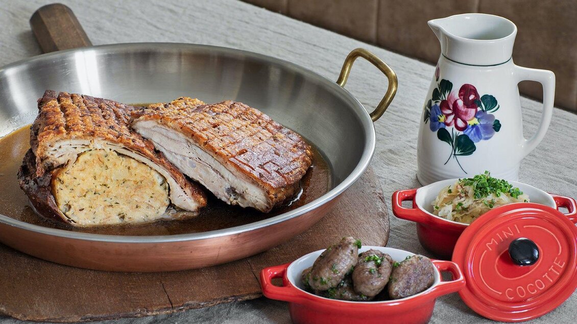 Ein leckerer Braten mit knuspriger Kruste und einer Füllung aus Kräutern in einer Bratpfanne. Dazu serviert sind kleine Würstchen und eine Kanne mit Sauce. | © GrafenWirt