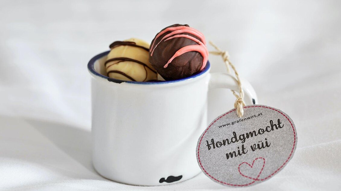 Eine kleine Tasse mit zwei handgemachten Pralinen, eine in Zartbitter- und eine in heller Schokolade. Ein Etikett mit der Aufschrift „Handgemacht mit viel Liebe“ hängt daran. | © GrafenWirt 
