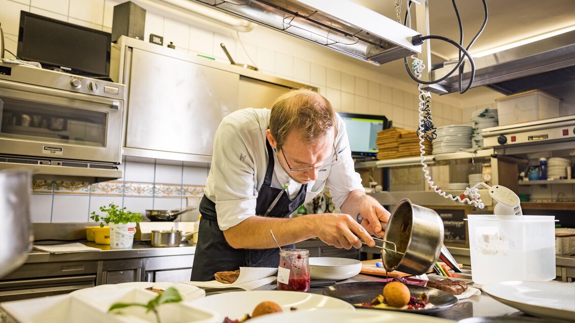 Ein Koch bereitet ein Gericht in einer modernen Küche vor. Er konzentriert sich auf das Anrichten auf einem weißen Teller. | © Netzwerk Kulinarik wildbild