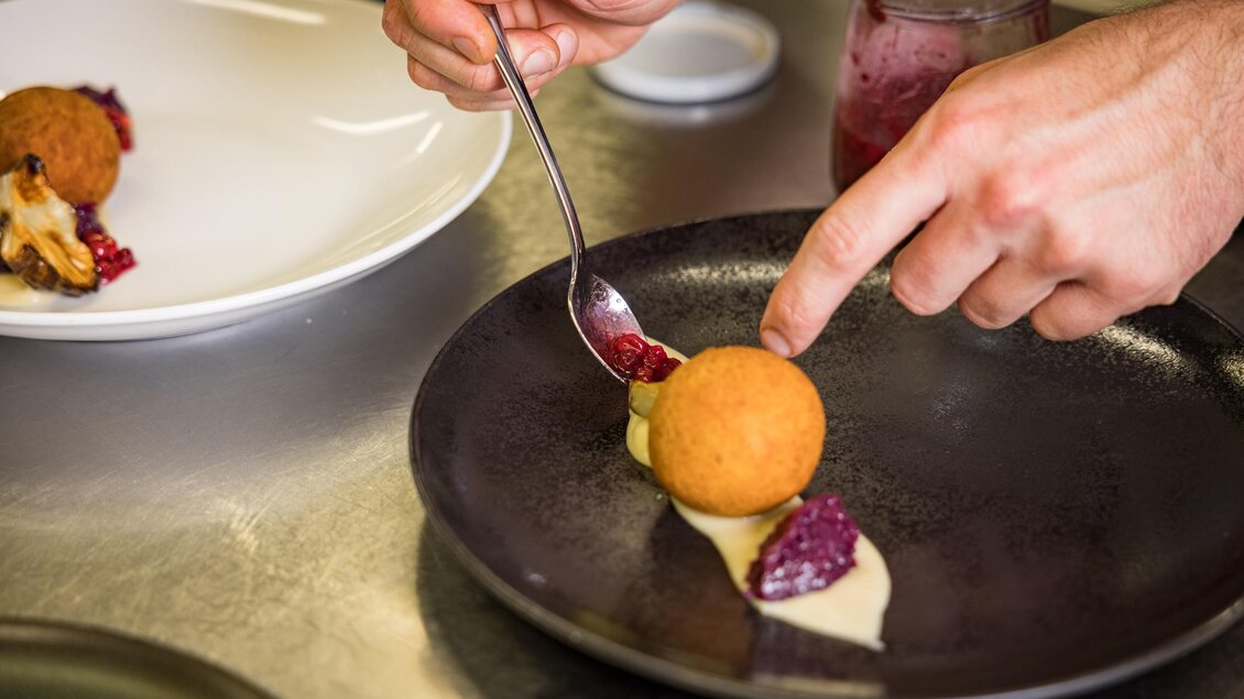Ein Koch dekoriert einen Teller mit einem runden Dessert und verschiedenen Saucen. Die Hände sind in Aktion, während das Gericht sorgfältig angerichtet wird. | © Netzwerk Kulinarik wildbild