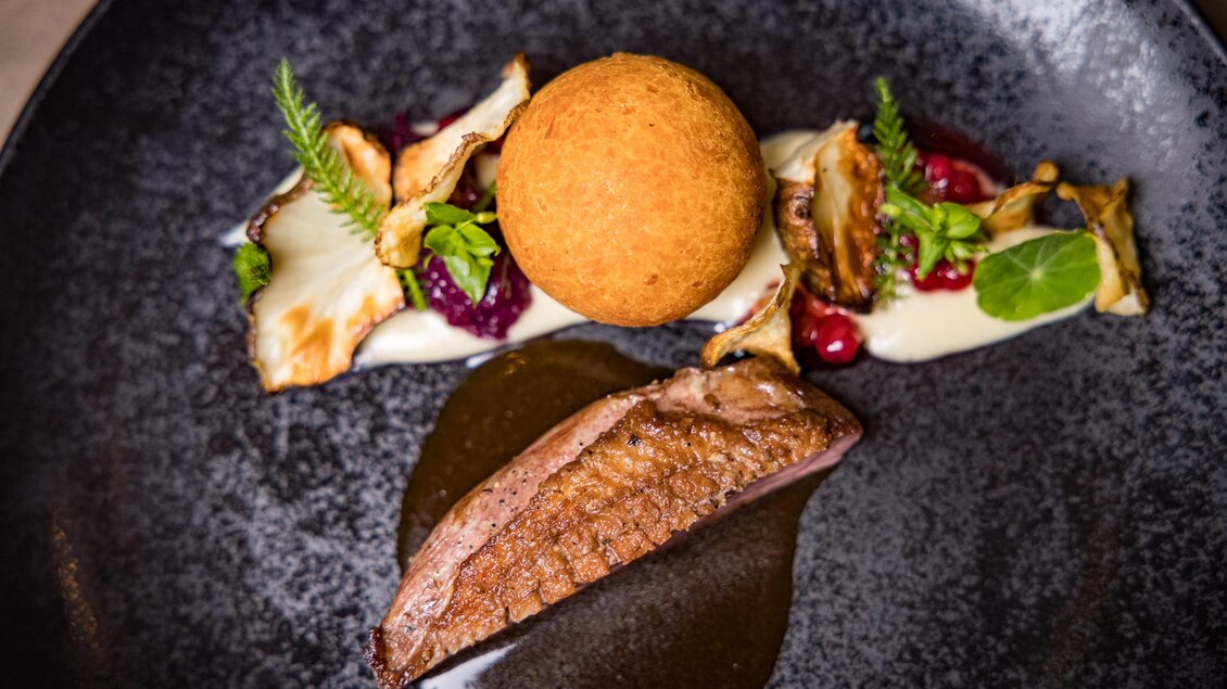 Ein kunstvoll angerichteter Teller mit gebratenem Fleisch, einer goldenen Kugel und verschiedenen Beilagen. Die Präsentation zeigt ein elegantes und modernes Gericht. | © Netzwerk Kulinarik wildbild