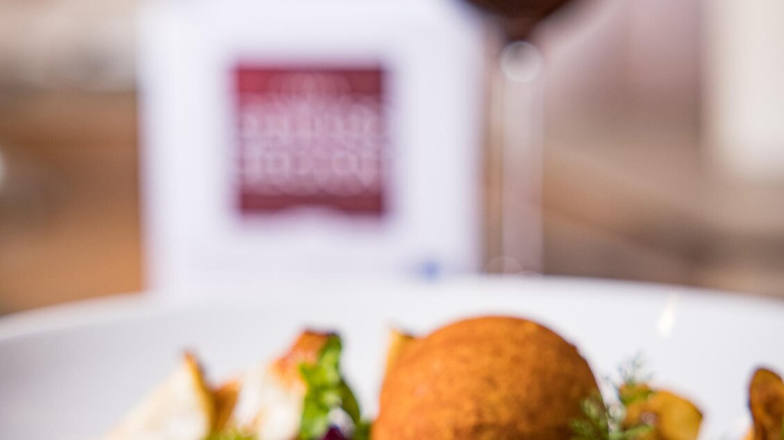 Ein köstliches Gericht mit perfekt gegartem Fleisch, einer goldbraunen Beilage und frischem Gemüse. Im Hintergrund ist ein Glas Rotwein zu sehen. | © Netzwerk Kulinarik wildbild