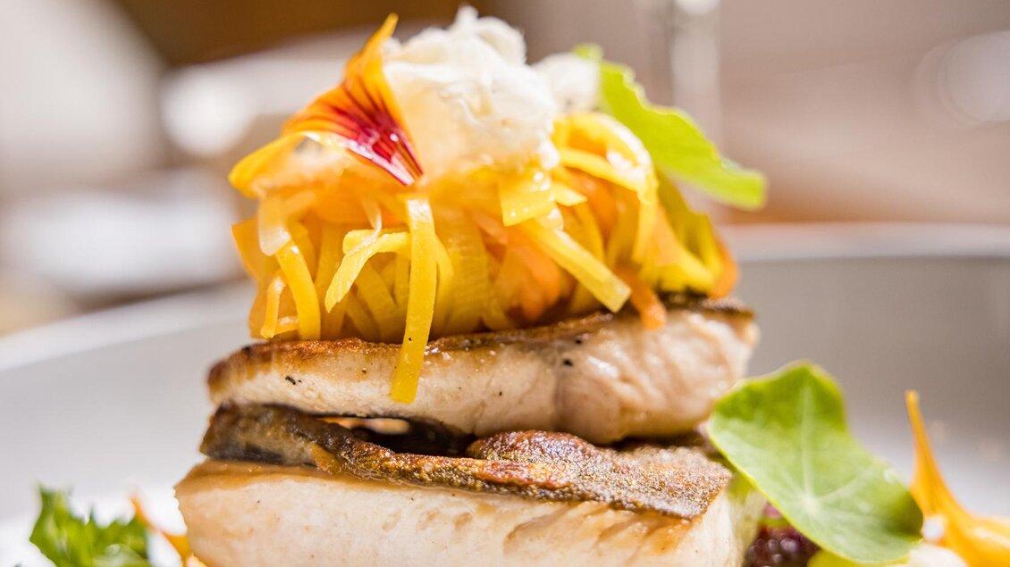 Ein schmackhaftes Gericht mit gebratenem Fisch, buntem Gemüse und einer cremigen Sauce. Im Hintergrund ist ein Glas Weißwein zu sehen. | © Netzwerk Kulinarik wildbild