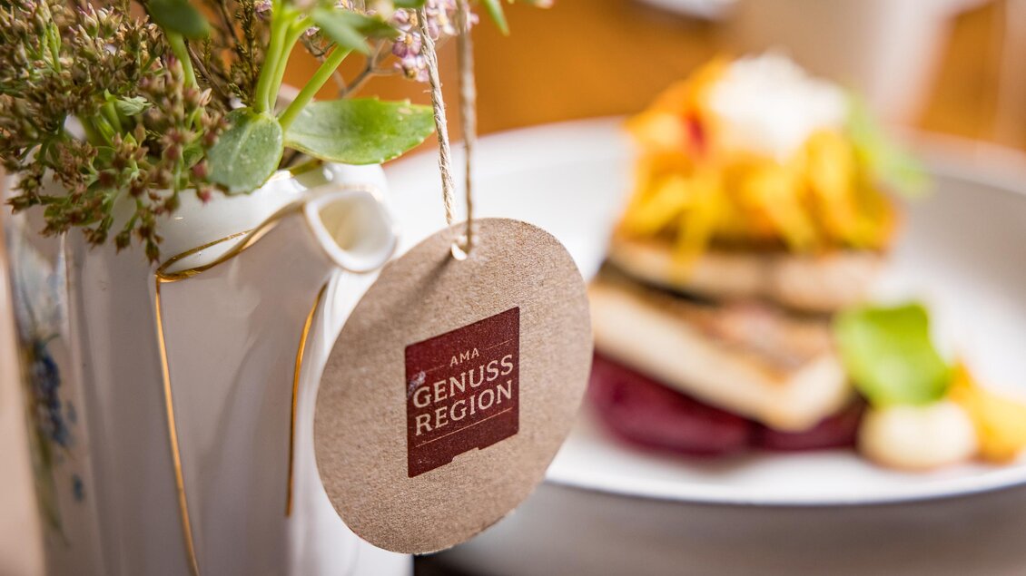 Ein hübsches Gefäß mit frischen Blumen und einem Etikett, das „Genussregion“ zeigt. Im Hintergrund ist ein Teller mit ansprechenden Speisen zu sehen. | © Netzwerk Kulinarik wildbild