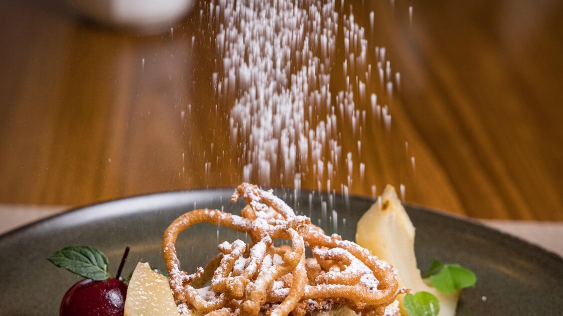 Ein kunstvoll angerichteter Teller mit einem Dessert, das mit Puderzucker bestreut wird. Neben dem Dessert liegen kleine Fruchtstücke und frische Kräuter. | © Netzwerk Kulinarik wildbild