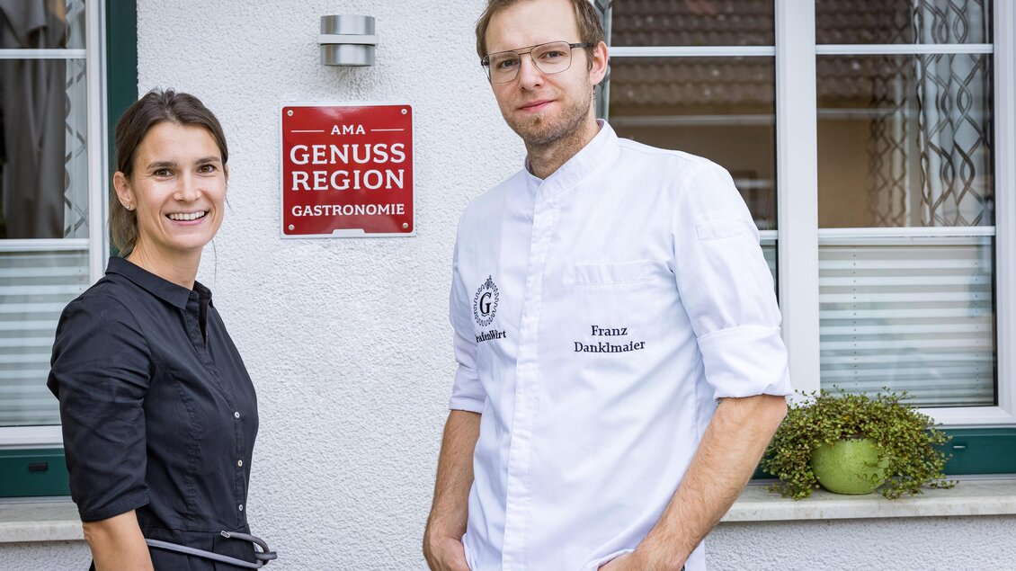 Ein Paar steht vor einem Schild, das auf die "Genuss Region" hinweist. Der Mann trägt eine Kochjacke und die Frau ein schwarzes Kleid. | © Netzwerk Kulinarik wildbild