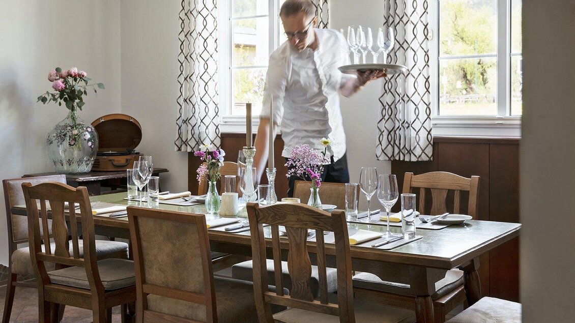 Ein elegantes Esszimmer mit einem Tisch, der für eine Mahlzeit gedeckt ist. Ein Kellner serviert Gläser und dekoriert den Tisch mit frischen Blumen. | © GrafenWirt