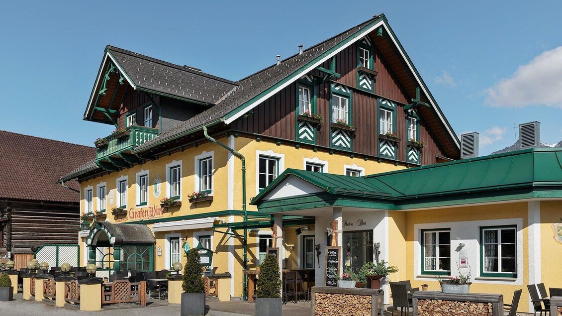 Ein traditionelles Gasthaus mit einer bunten Fassade. Vor dem Gebäude befinden sich Sitzmöglichkeiten im Freien und hübsche Pflanzen. | © GrafenWirt