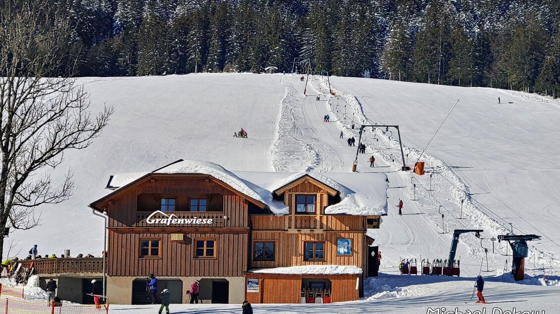 Ein hübsches Holzhaus in einem schneebedeckten Gebiet. Im Hintergrund sind Skifahrer und ein Schlepplift zu sehen. | © Melanie Fritz_Steirisch Sche