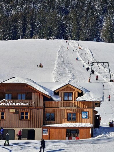 Ein hübsches Holzhaus in einem schneebedeckten Gebiet. Im Hintergrund sind Skifahrer und ein Schlepplift zu sehen. | © Melanie Fritz_Steirisch Sche