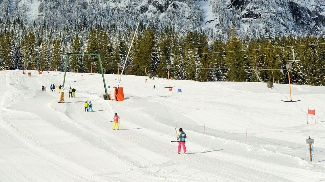 Eine schneebedeckte Skipiste mit Skifahrern und einem Skilift im Hintergrund. Im Wald hinter den Pisten sind hohe, grüne Bäume zu sehen. | © Thomas Sölkner