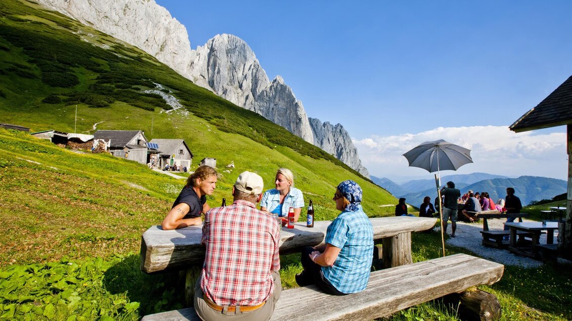 Eine Gruppe von Personen sitzt an einem Holztisch in den Bergen. Im Hintergrund sind grüne Wiesen und beeindruckende Felsen zu sehen.
