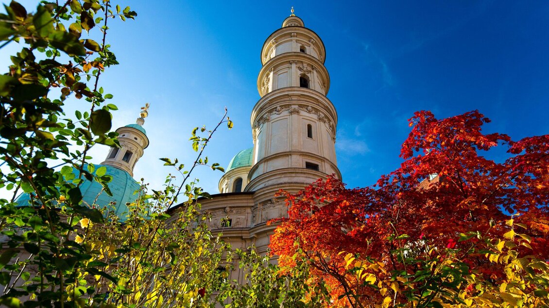 Eine beeindruckende Kirchturmarchitektur umgeben von buntem Herbstlaub. Der klare blaue Himmel verstärkt die Schönheit der Szene. | © Graz Tourismus - Harry Schiffer