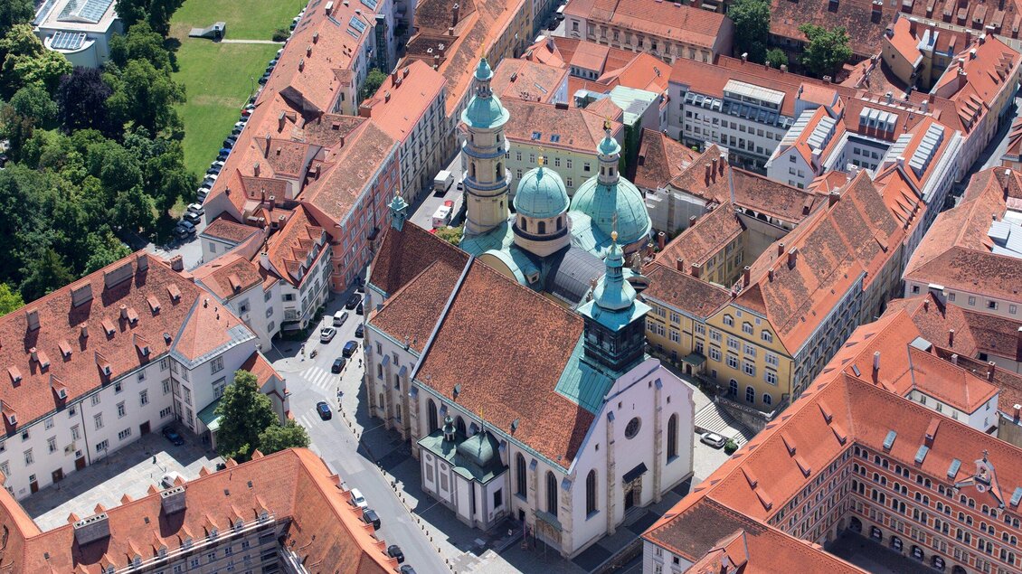 Eine beeindruckende Ansicht einer Stadt mit roten Dächern und einer markanten Kirche mit grünen Kuppeln. Das Bild zeigt die architektonische Schönheit und die dichte Bebauung der Umgebung. | © Graz Tourismus - Harry Schiffer