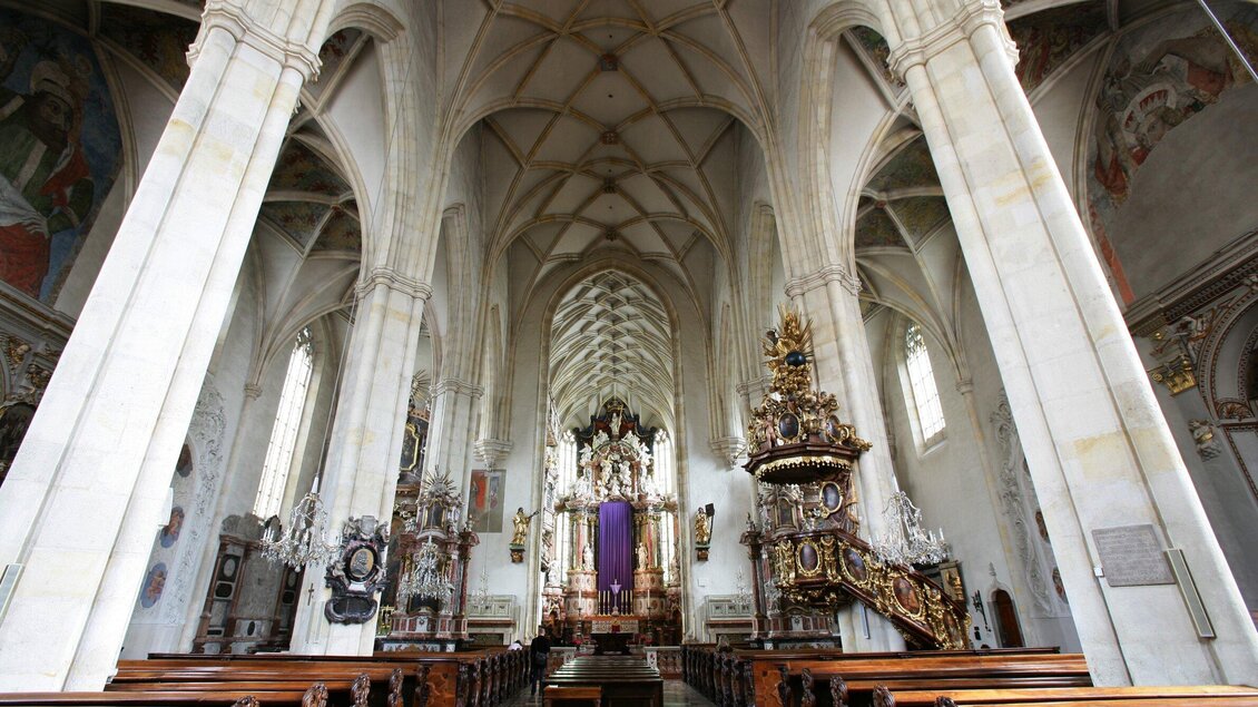 Ein prächtiger Innenraum einer Kirche mit hohen, gewölbten Decken und beeindruckenden Säulen. Der Altar ist kunstvoll verziert und ein violettes Tuch hängt von der Empore. | © Graz Tourismus - Harry Schiffer