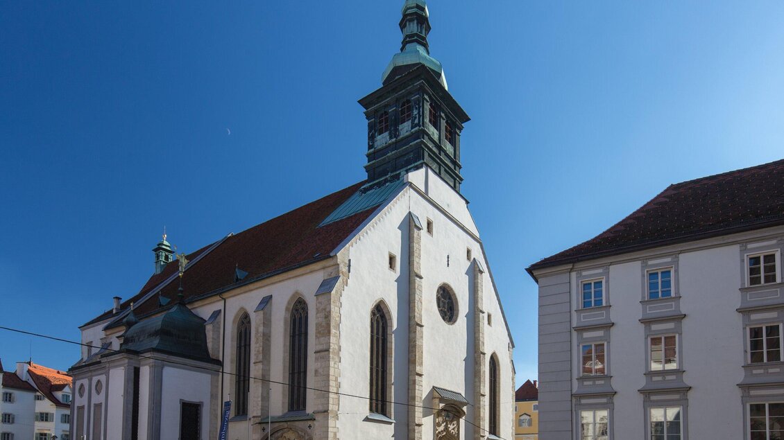 Eine historische Kirche mit einem markanten Turm und weißer Fassade. Der blaue Himmel sorgt für eine klare und helle Atmosphäre. | © Graz Tourismus - Harry Schiffer