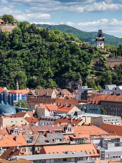 Eine malerische Stadtansicht mit roten Dächern und üppigem Grün. Im Hintergrund ist eine beeindruckende Festung auf einem Hügel zu sehen. | © Graz Tourismus - Harry Schiffer