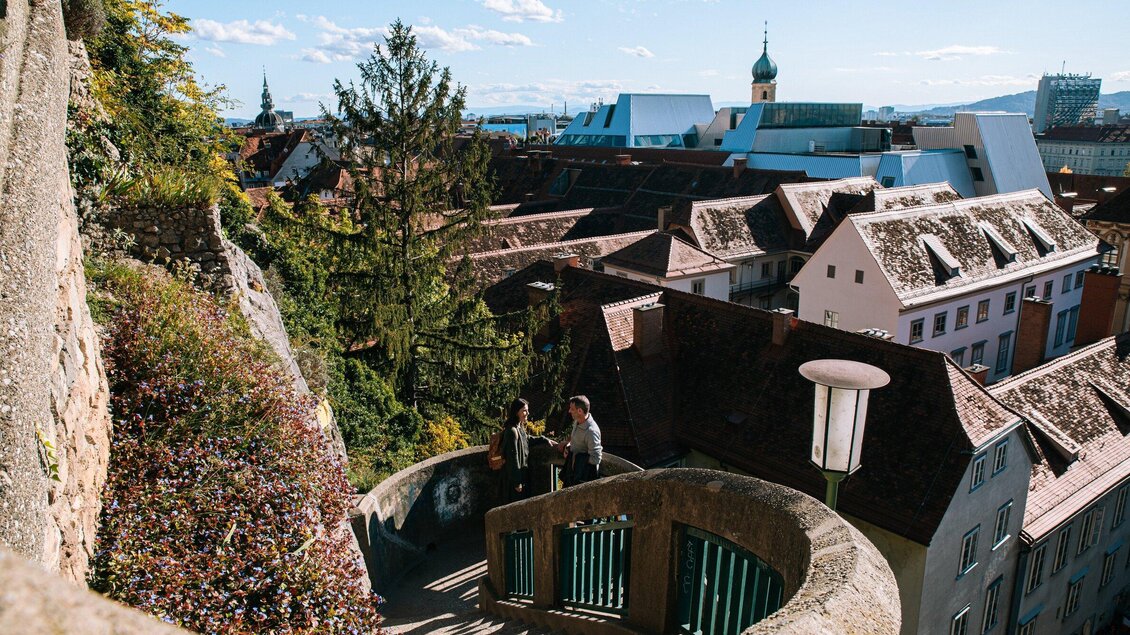 Eine malerische Aussicht auf die Dächer einer Stadt mit charmanten Gebäuden. Im Vordergrund stehen zwei Personen auf einer geschwungenen Treppe. | © Graz Tourismus