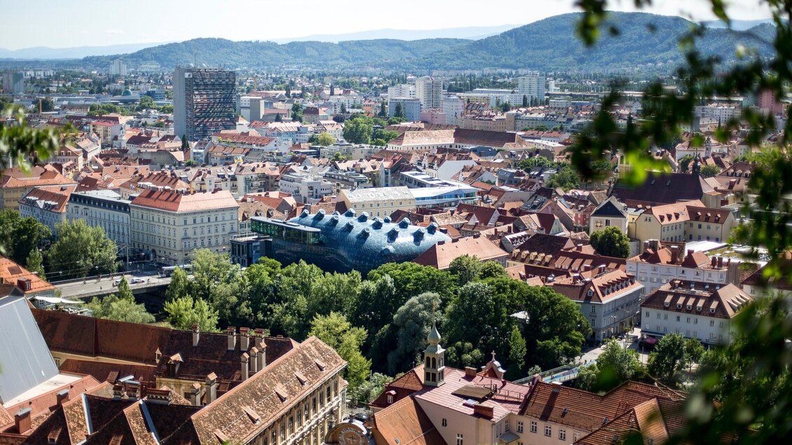 Eine beeindruckende Stadtansicht mit roten Dächern und grünen Bäumen. Im Hintergrund sind moderne Gebäude und sanfte Hügel zu sehen. | © Graz Tourismus - Tom Lamm
