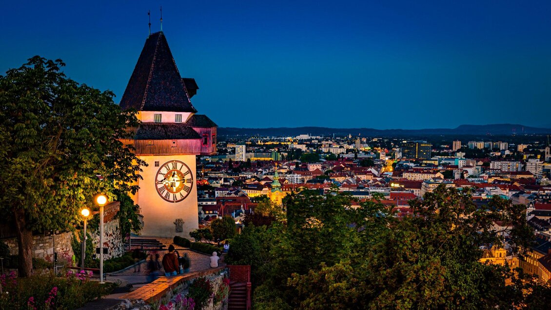 Ein historischer Turm mit einer großen Uhr bei Nacht. Die Stadt leuchtet unter dem klaren Himmel im Hintergrund. | © Graz Tourismus - Werner Krug