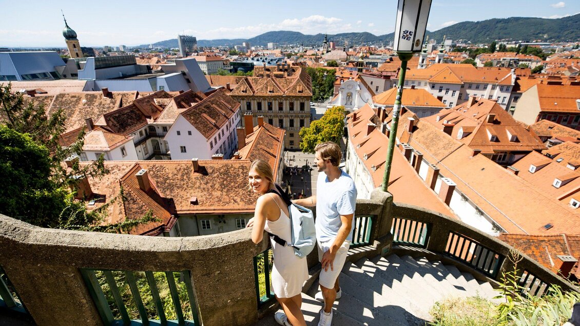Ein Blick auf eine malerische Stadt mit roten Dächern und grünen Hügeln im Hintergrund. Zwei Personen stehen auf einer Treppe und genießen die Aussicht. | © Graz Tourismus - Tom Lamm
