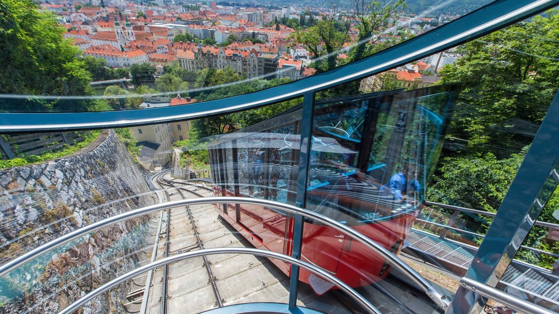 Eine beeindruckende Aussicht auf die Stadt von einer Standseilbahn. Die rot lackierte Bahn fährt eine steile Strecke hinunter, umgeben von Bäumen. | © Graz Tourismus - Harry Schiffer