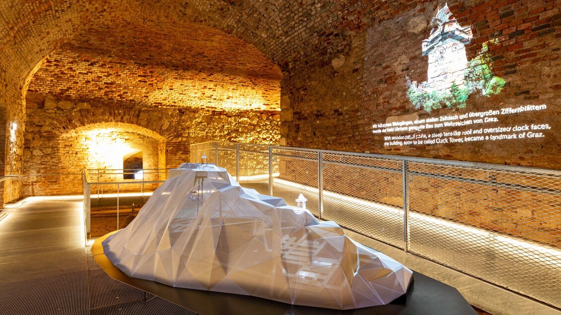 Ein moderner, geometrischer Kunstgegenstand steht in einem historischen Raum mit Ziegelmauern. An der Wand sind projizierte Informationen und Bilder zu sehen. | © Graz Tourismus - Harry Schiffer