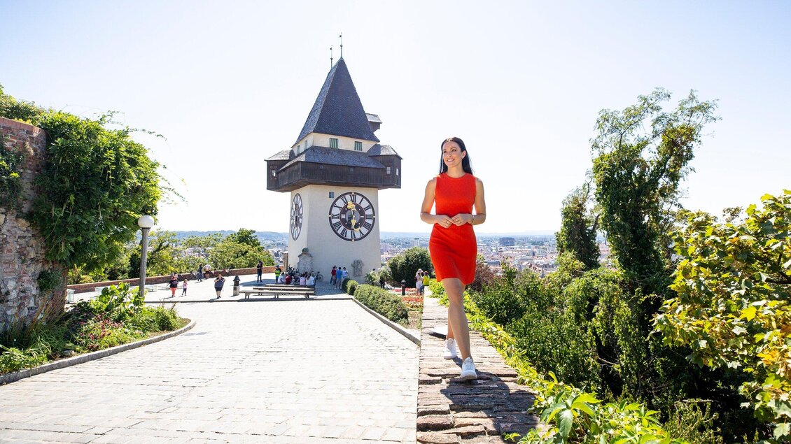 Eine Frau in einem roten Kleid geht einen Weg entlang, der zu einem historischen Turm führt. Im Hintergrund sind Menschen und eine schöne Landschaft zu sehen. | © Graz Tourismus - Tom Lamm