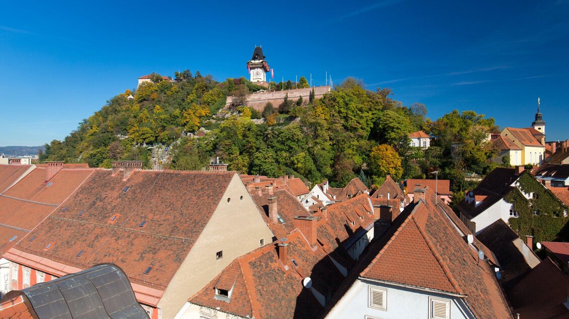 Eine malerische Ansicht von roten Dächern und einem Hügel mit einer Burg im Hintergrund. Der klare blaue Himmel ergänzt die schöne Landschaft. | © Graz Tourismus - Harry Schiffer