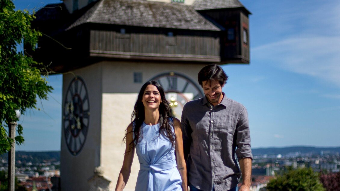 Ein lächelndes Paar spaziert in der Nähe eines historischen Turms an einem sonnigen Tag. Die klare Himmel und die grüne Umgebung schaffen eine einladende Atmosphäre. | © Graz Tourismus - Tom Lamm