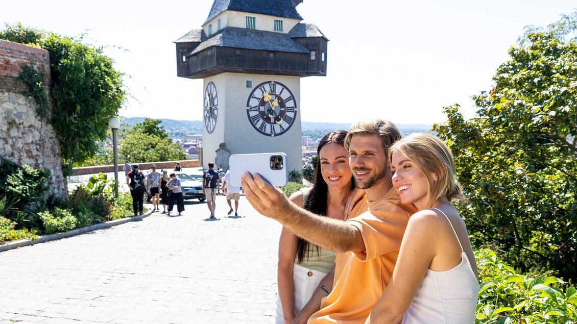 Drei Freunde machen ein Selfie vor einem historischen Turm. Im Hintergrund sind grüne Bäume und eine schöne Aussicht zu sehen. | © Graz Tourismus - Tom Lamm